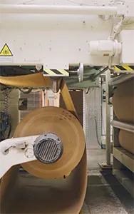 A large roll of brown paper or material is being unwound by an industrial machine in a factory. Above the roll, yellow and black striped warning tape is visible on a white metal structure, and a yellow triangular warning sign is also present.