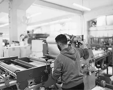 A black and white photo shows a person from behind, wearing a hoodie, operating a large industrial printing or manufacturing machine. Various rollers and mechanical parts are visible on the machine, and a blurred factory interior is in the background.