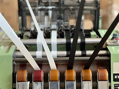 A close-up view of a weaving machine with multiple white, black, and light-colored ribbons or tapes being fed through rollers. Below the tapes, a row of brown and red spools are visible. Part of a label on the machine reads "KYANG".