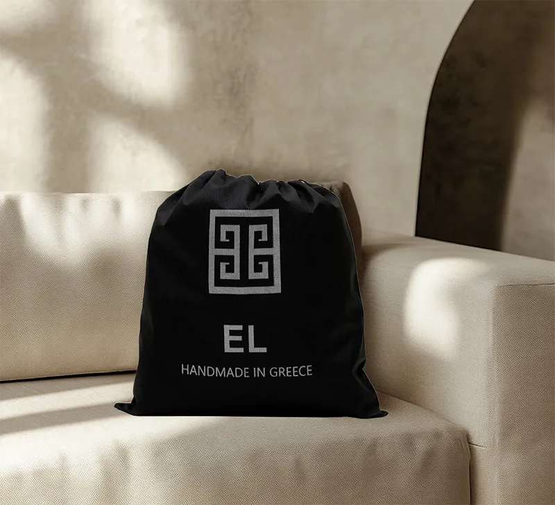 A black drawstring dust bag with a silver logo is positioned upright on a light beige sofa. The logo consists of an intricate, square, maze-like or Greek key pattern above the letters "EL" in a bold, sans-serif font, and "HANDMADE IN GREECE" in a smaller font below. The sofa has clean lines and light-colored upholstery, and the background shows a textured wall with subtle shadows, possibly from natural light.