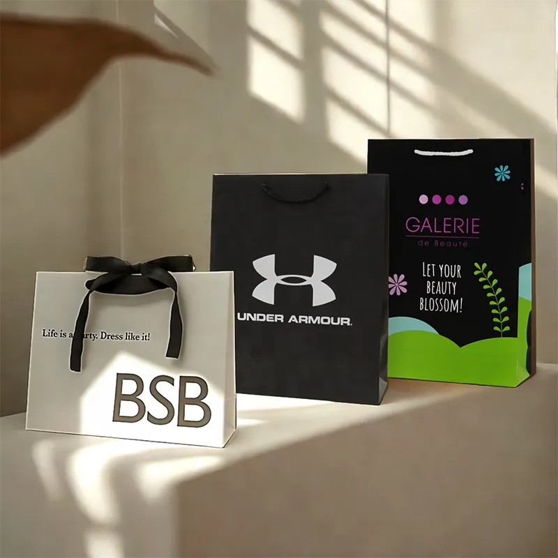 Three distinct paper shopping bags are displayed, featuring a white bag with a black ribbon and "BSB" branding, a black bag with the "Under Armour" logo, and a black bag with colorful "GALERIE de Beauté" branding and floral designs.