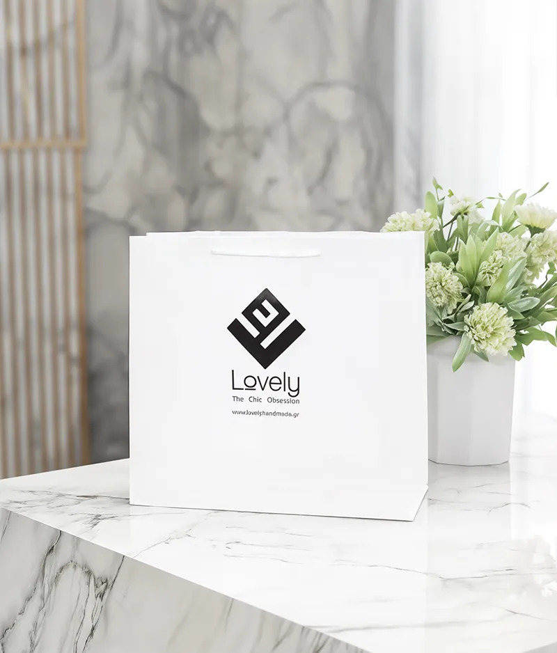 A clean, white paper shopping bag with black text and a logo is centered on a white marble-patterned surface. The bag has white cord handles. The logo is a stylized, angular design, resembling an "L" or an abstract shape. Below the logo, the text reads "Lovely" in a script font, followed by "The Chic Obsession" in a smaller, sans-serif font, and finally "www.lovelyishandmade.gr" in an even smaller font. 