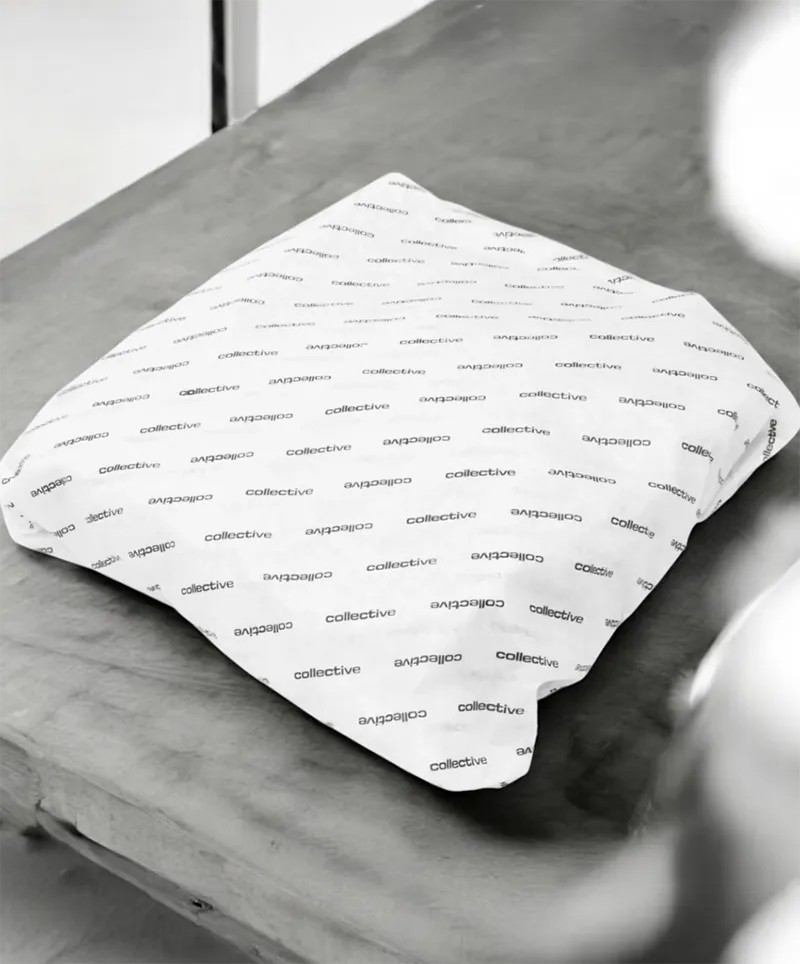 A monochromatic image shows a rectangular item, likely a soft good or apparel, neatly wrapped in white tissue paper. The tissue paper is uniformly patterned with the word "collective" repeated multiple times in a small, black, sans-serif font. The wrapped item rests on a surface that appears to be made of wood or a similar textured material. The lighting in the image suggests a soft, diffused light source, highlighting the texture of the wrapping paper and the surface it rests on.