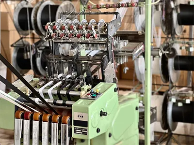 A close-up of a textile weaving machine with various white and black ribbons or tapes being woven. Numerous spools of thread are visible on the machine's frame and in the background, which is blurred. A green control panel with "KY" visible is part of the machine.