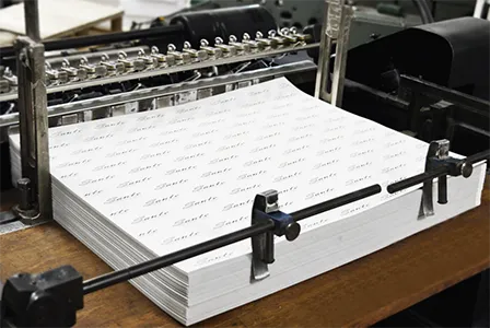 A stack of printed papers, featuring a dark cover with "HEIDELBERG SPEEDMASTER" and "OUTLET" text, rests in the output tray of a large, dark gray printing press. In the blurred background, more industrial printing machinery is visible.