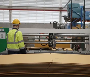 A person wearing a yellow hard hat and a high-visibility yellow and black jacket stands with their back to the viewer, looking at large industrial machinery. In the foreground, a stack of flat, pre-cut cardboard pieces is ready to be processed. The background shows a factory setting with shelves and equipment.