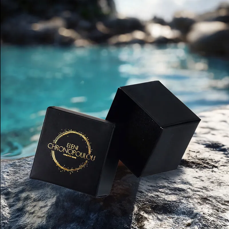 Two small, square black jewelry boxes are placed on a rough, dark gray rock surface. One box is closed, displaying a gold circular logo with "ELENI CHRONOPOULOU" and additional smaller Greek text within. The other box is open, revealing its black interior, and is slightly tilted. In the blurred background, a body of turquoise or blue water is visible, suggesting a natural, possibly coastal or lakeside, setting.
