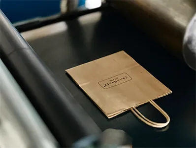 A small, flat brown paper bag with string handles and a black stamped logo that reads "C A S T I N G R E A T" is centered on a dark, reflective surface next to metallic machinery parts.