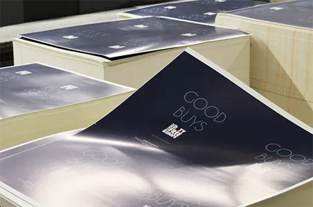 Stacks of freshly printed brochures or magazines are seen, with the top sheet featuring a dark background and white text that reads "GOOD BUYS" and "SAVE UP TO 70% FREE SHOPS". The light reflects off the glossy surface of the prints.