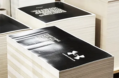 Stacks of printed materials with a prominent black cover featuring the Under Armour logo and the text "EVERYTHING ENGINEERED TO MAKE YOU BETTER." in white. The pages beneath the cover appear to be white.